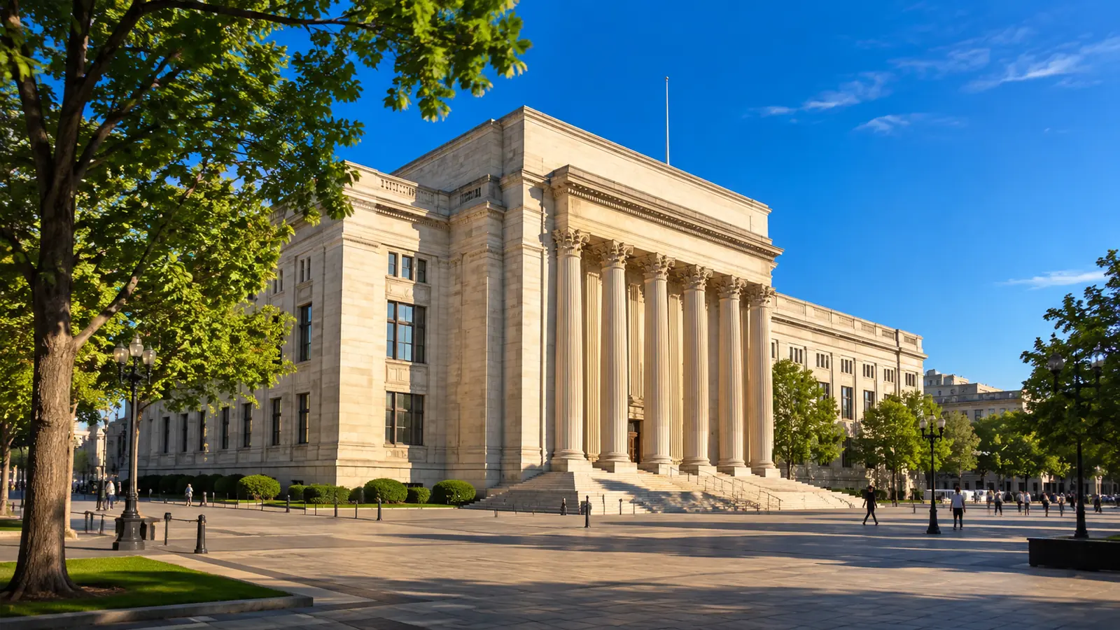 A stone central bank building across a sunlit plaza on a clear summer morning, with street trees and distant pedestrians