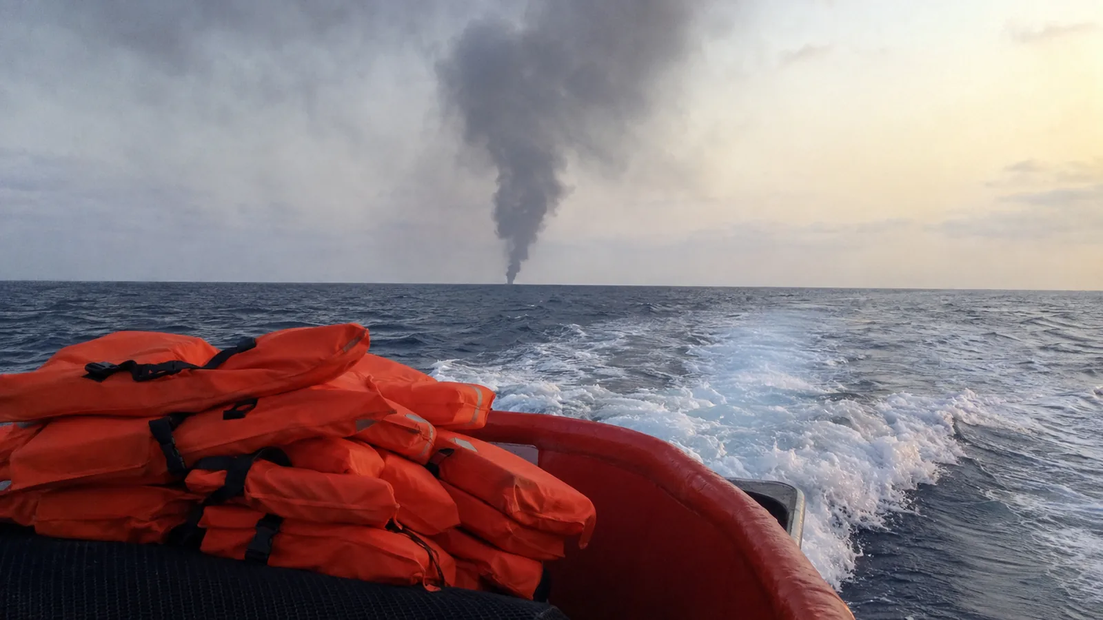 Smoke on the horizon seen from a rescue boat's deck, life jackets stacked ready in the foreground