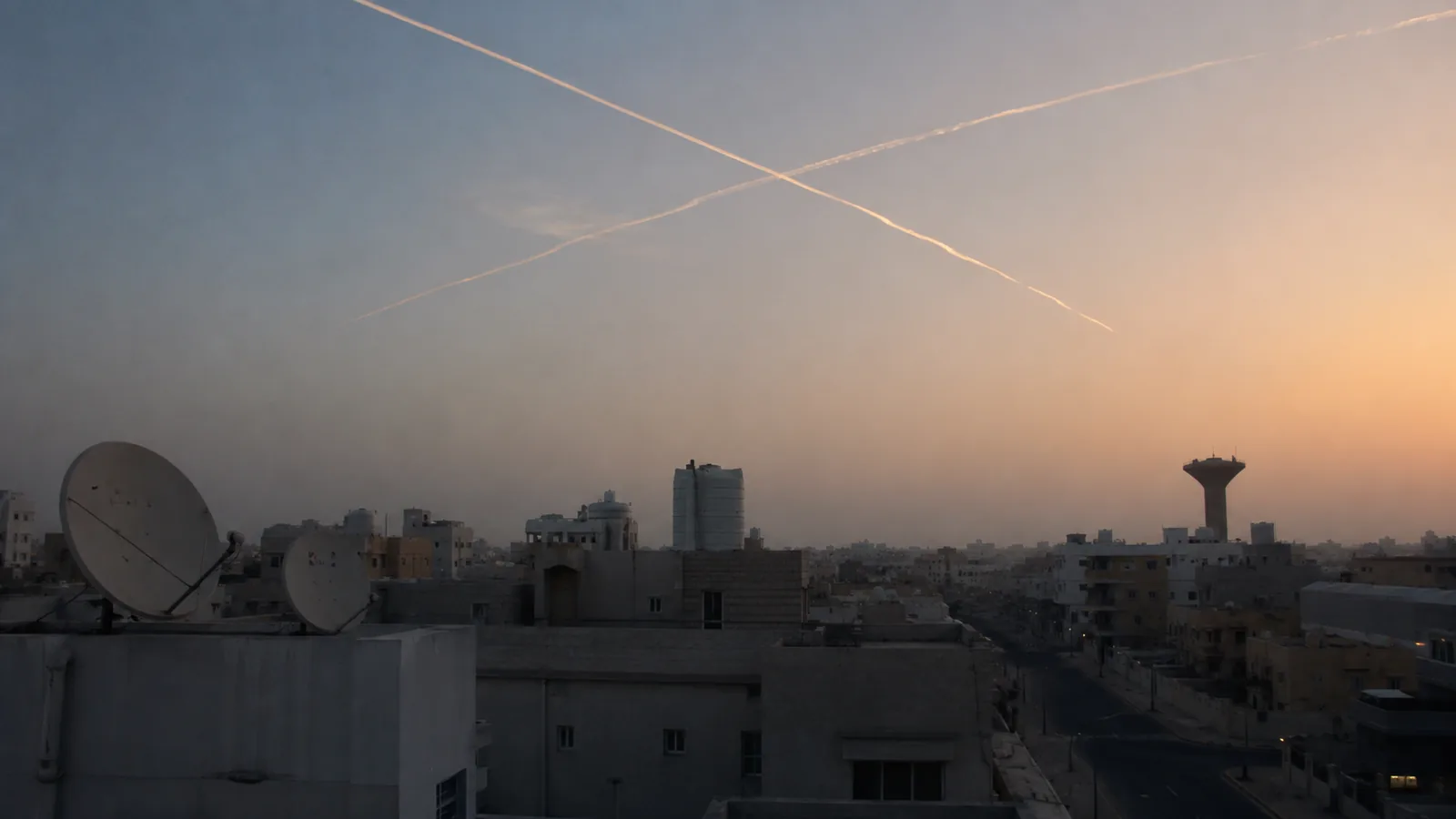Two interception trails crossing a pale dawn sky above a Gulf city's rooftops