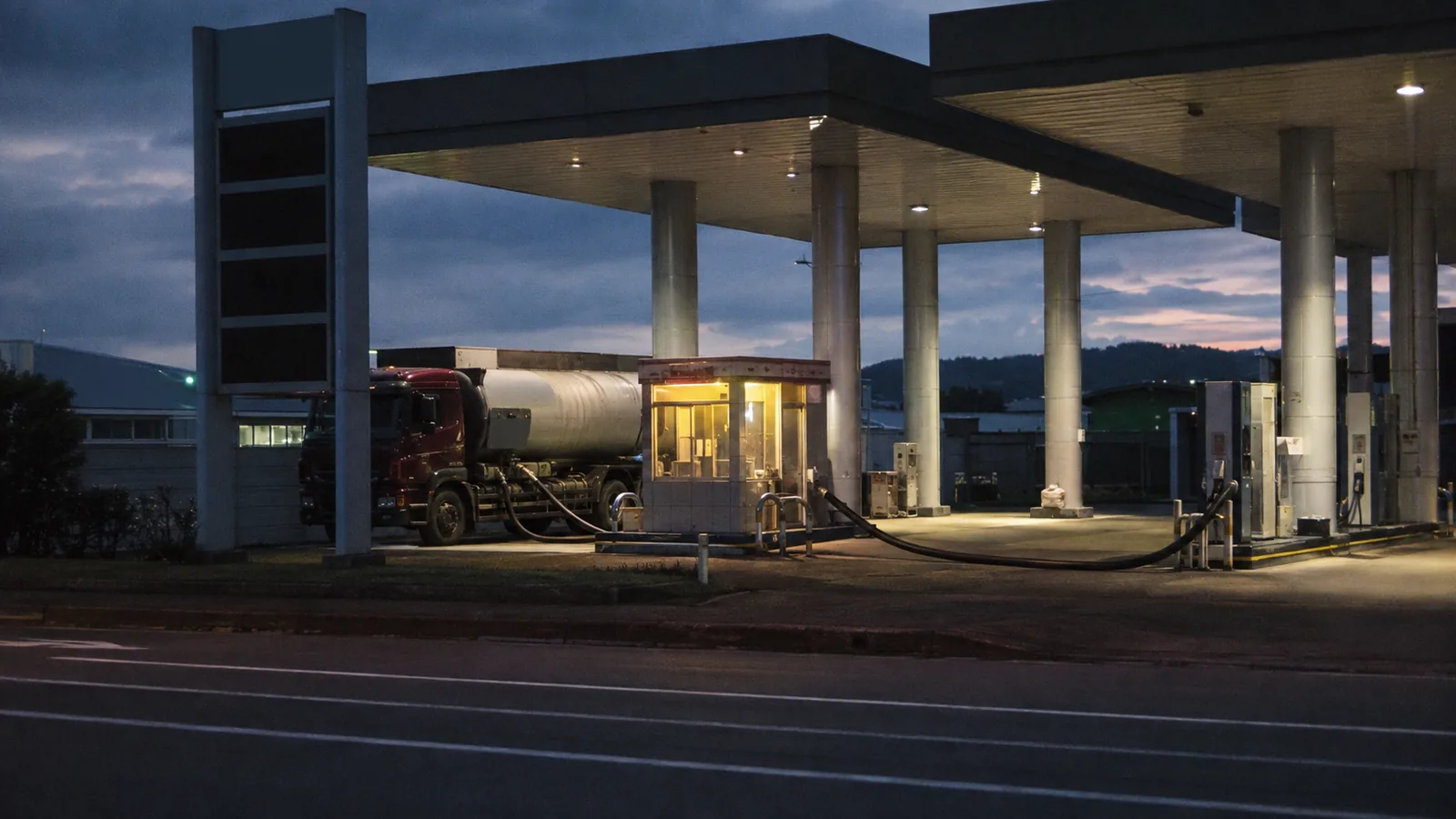 A tanker truck unloading into a gas station's underground tanks at dusk, price display dark