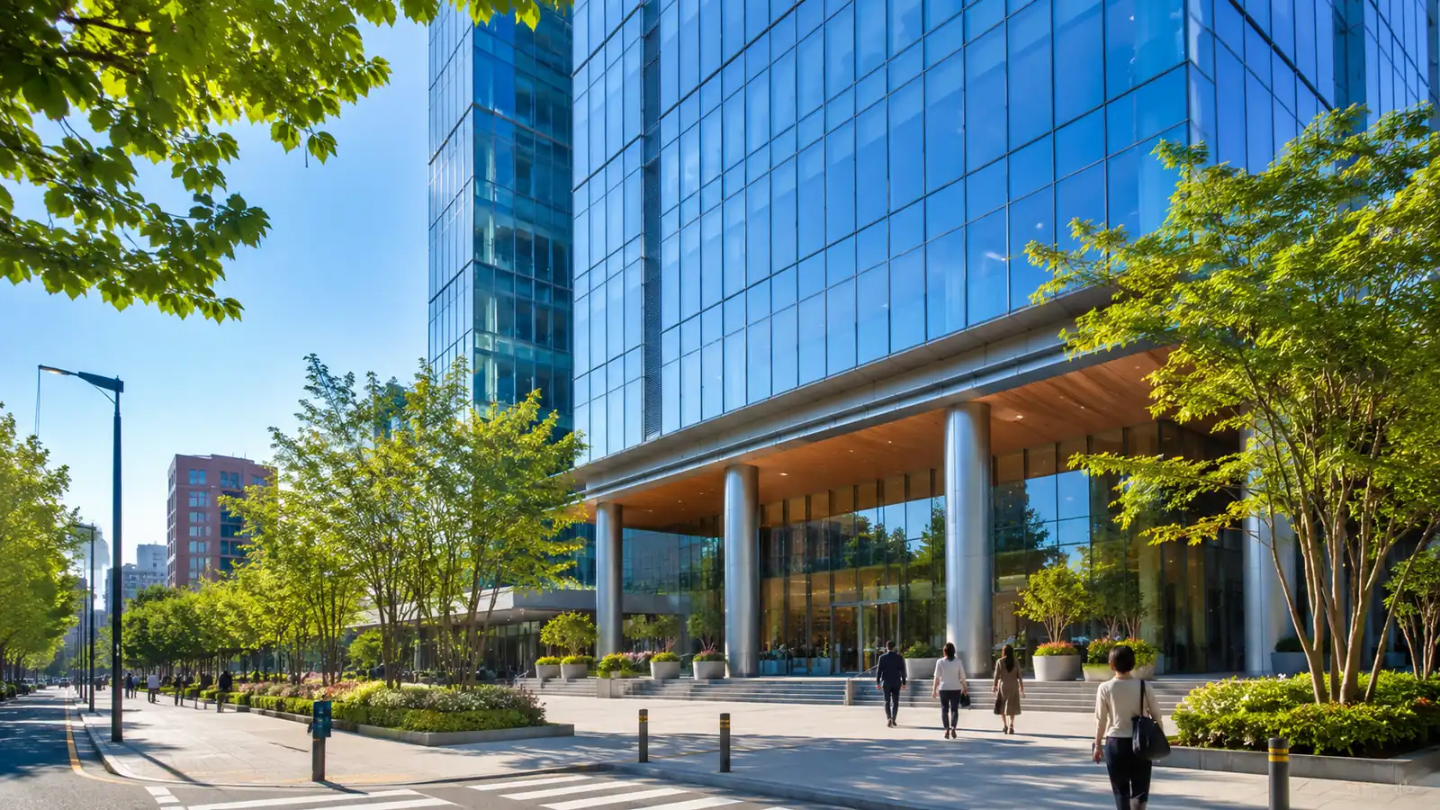 A modern glass office tower seen from a sunny street corner in mid-morning, with green street trees along the sidewalk