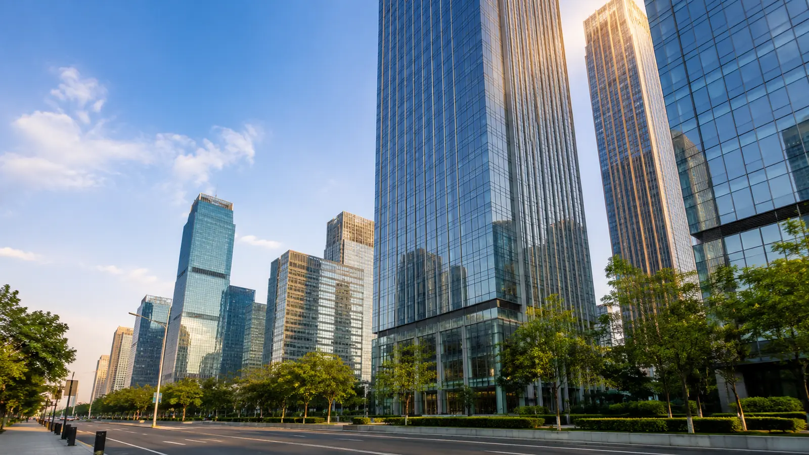 Glass office towers in a financial district with morning sunlight on the upper floors and trees lining the sidewalk