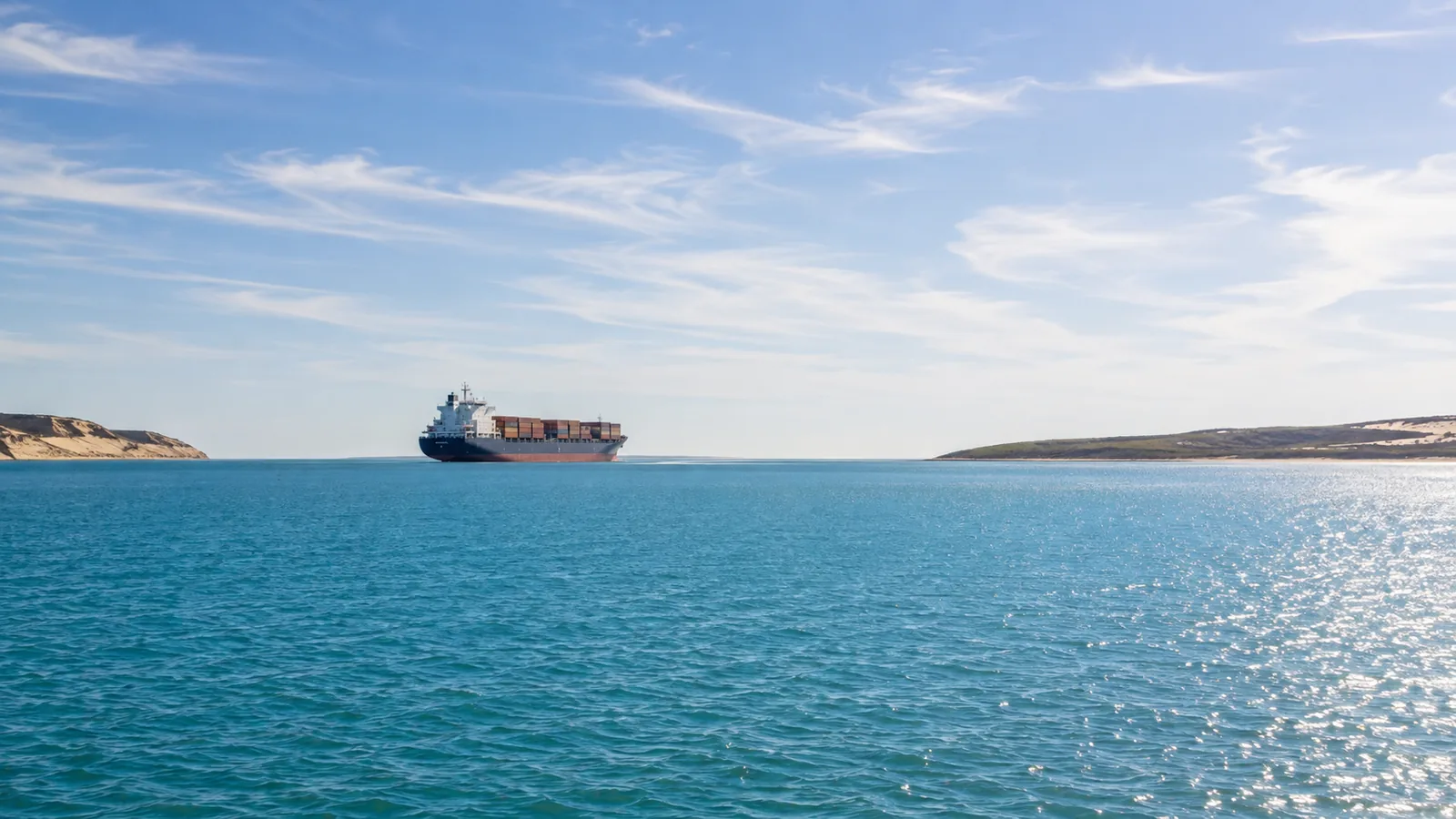 A large cargo ship crossing calm blue-green water at midday with low sandy headlands on the horizon