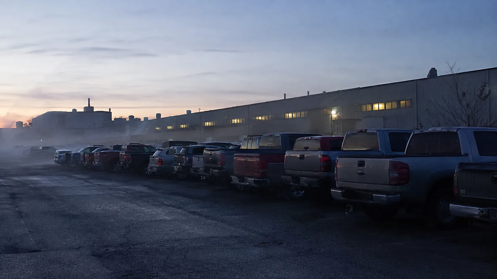 A factory lot at dawn shift change — rows of pickup trucks in the mist
