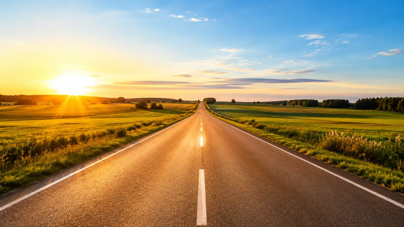 A long straight road stretching to the horizon under a clear morning sky, green fields on both sides